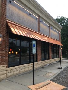 A long row of storefront windows set in a brick exterior wall and shaded by a rust-colored standing seam awning 