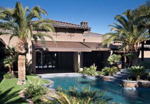 Reddish-tan stucco home's backyard with a rust-colored retractable awning covering a patio/pool deck, swimming pool and lush green trees and grass.
